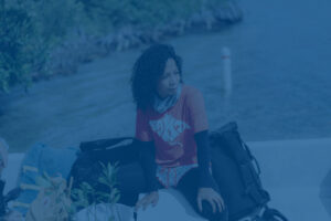 A woman on a boat sits with a bucket of mangrove saplings in front of her.